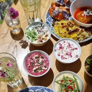 Sunlit wooden table with bowls of yogurt dips garnished with rose petals, a plate of saffron rice with stew, and glasses of iced drinks