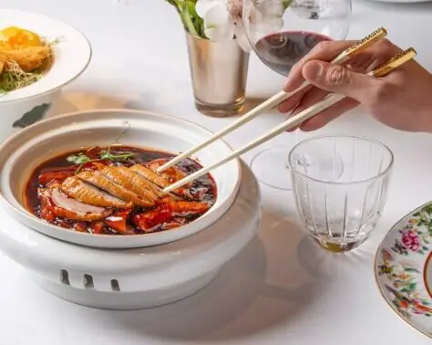 Sliced roast duck in a red broth in a round bowl, chopsticks lifting a piece, wine glass and place setting nearby