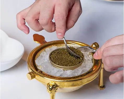 Hands spooning caviar from a tin set in ice in a gold serving dish