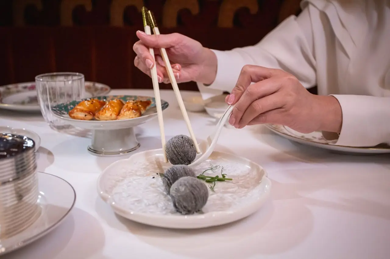 Chopsticks and a spoon lifting a dark grey dumpling from a plate of three, pastries visible on a stand in the background