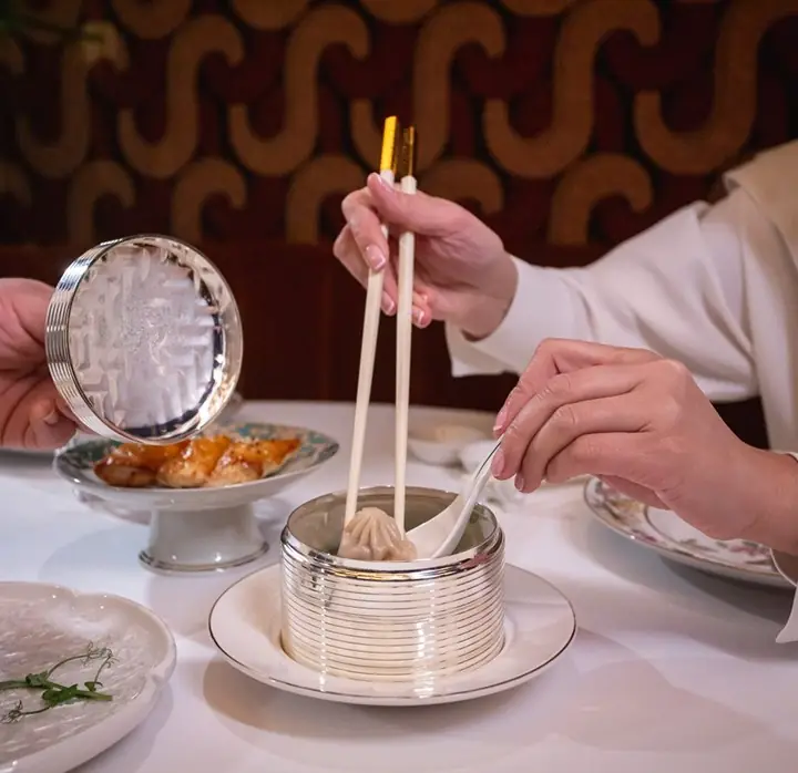 Hands using chopsticks and a spoon to lift a soup dumpling from a silver-lidded steamer