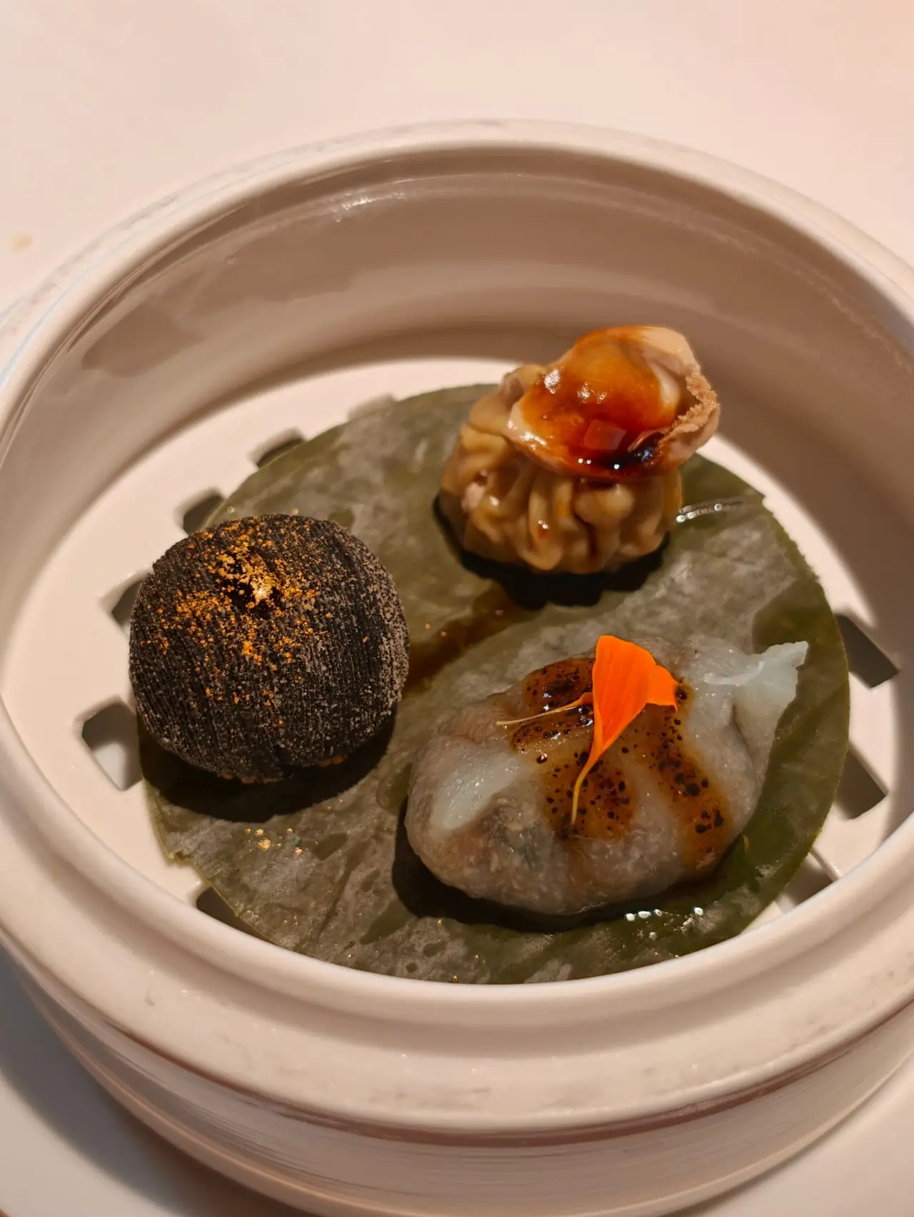 Three dim sum dumplings in a bamboo-style steamer, including a black sesame ball and a shrimp dumpling with sauce