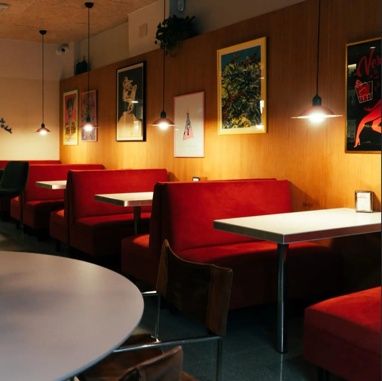 Dim interior with red upholstered booths, white tables, framed vintage posters on wood-panelled walls and red dome pendant lamps