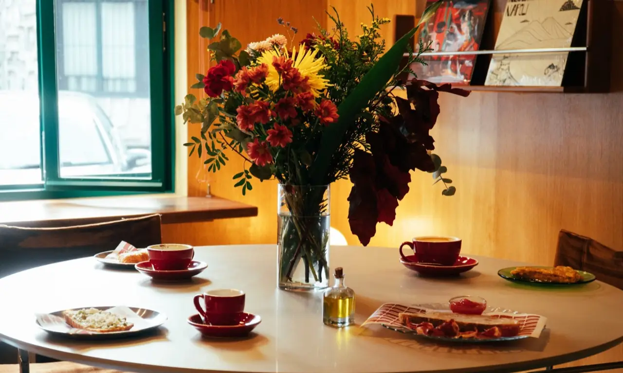 Round white table set with red espresso cups, tostadas, a bottle of oil and a large flower bouquet in a glass vase by a window