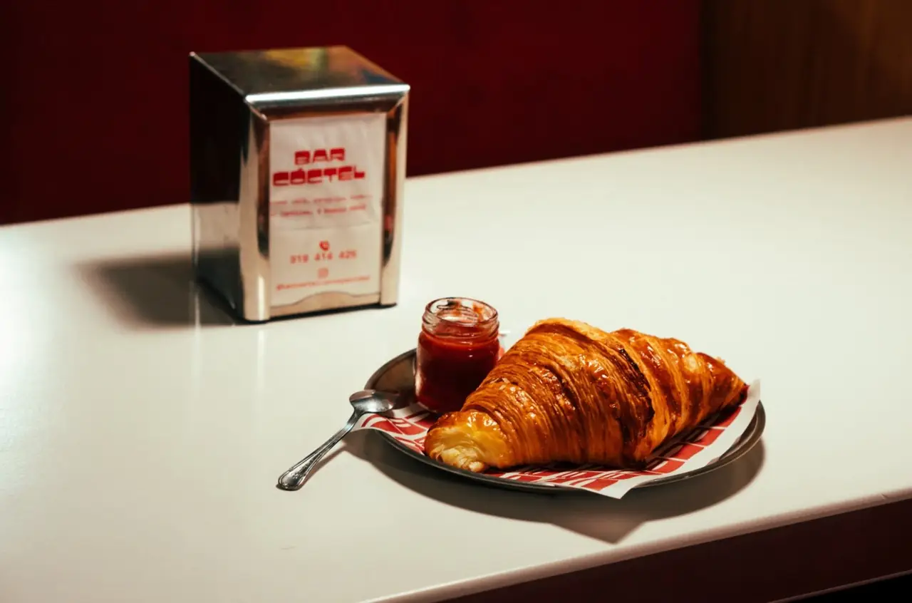 Glazed croissant on a metal plate with a small jar of jam and spoon, beside a chrome napkin dispenser branded Bar Coctel