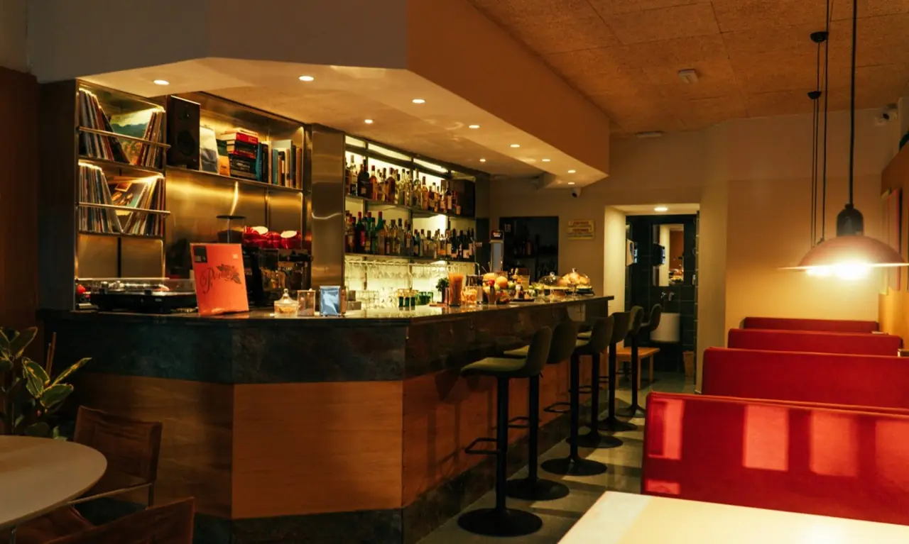 Bar interior with a stone-topped counter, backlit spirits shelf, vinyl records and books, black bar stools and red booths