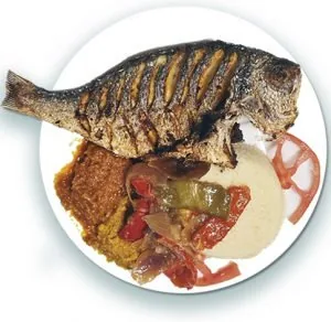A whole grilled fish served over rice with stewed peppers and onions, a pale puree and slices of tomato