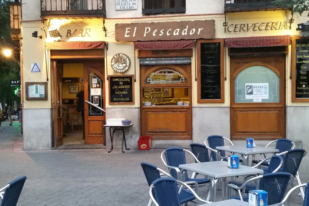 The corner frontage of Bar El Pescador Cerveceria at dusk, blackboards advertising raciones and bocadillos beside a hand-lettered 'Especialidad en Calamares' sign, blue wicker terrace chairs out front