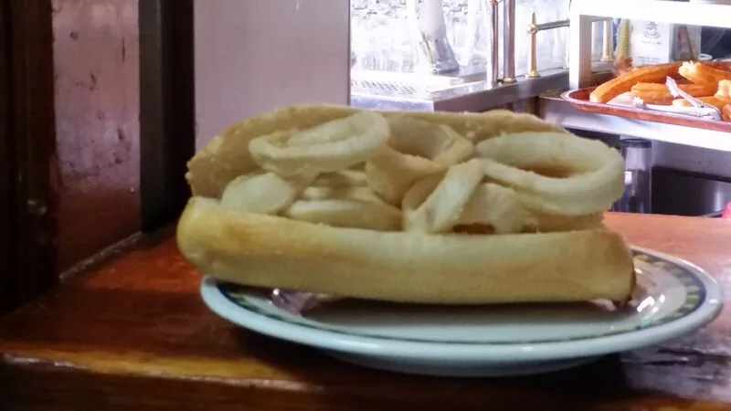 A bocadillo de calamares, a baguette packed with fried squid rings, on a small plate on the wooden bar top, beer taps and a tray of churros behind