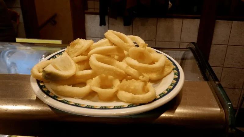 A plate piled with battered fried calamari rings and a lemon wedge, resting on the bar's metal counter rail