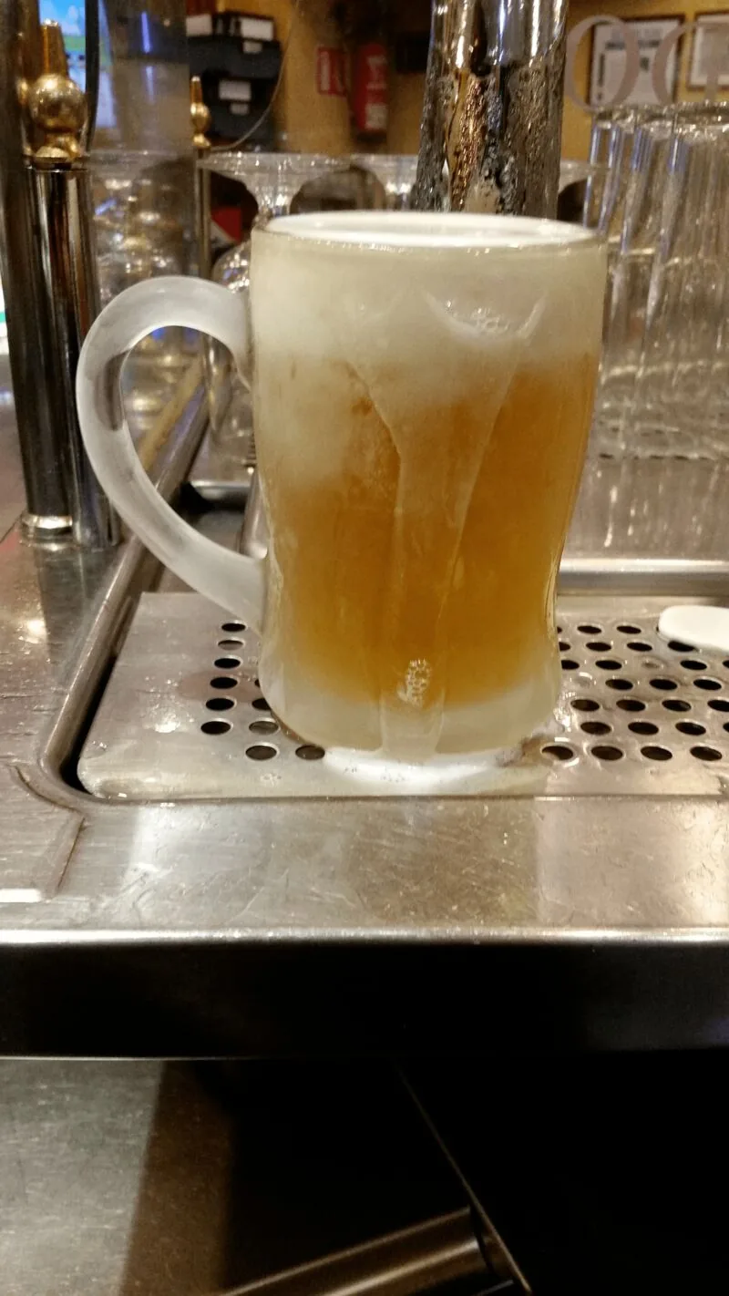 A frosted glass tankard of beer with a thick head, just poured and sitting on the drip tray beneath the taps