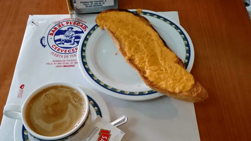 A toasted half-baguette spread with grated tomato beside a cortado in a small cup, on a paper placemat printed with the Bar El Pescador Cerveceria logo