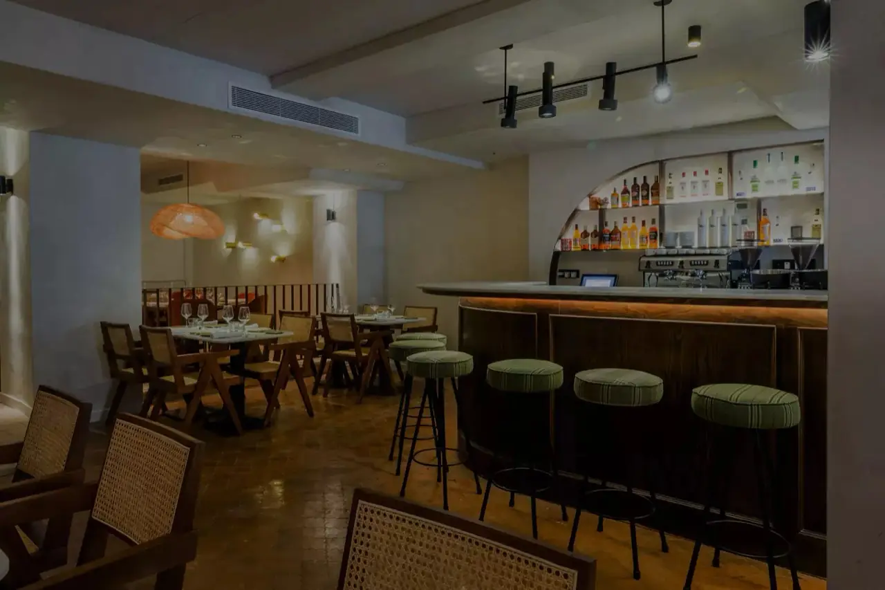 Dining room with wood-and-cane chairs, a curved dark-wood bar topped in pale stone, green upholstered bar stools and a lit back bar of spirit bottles