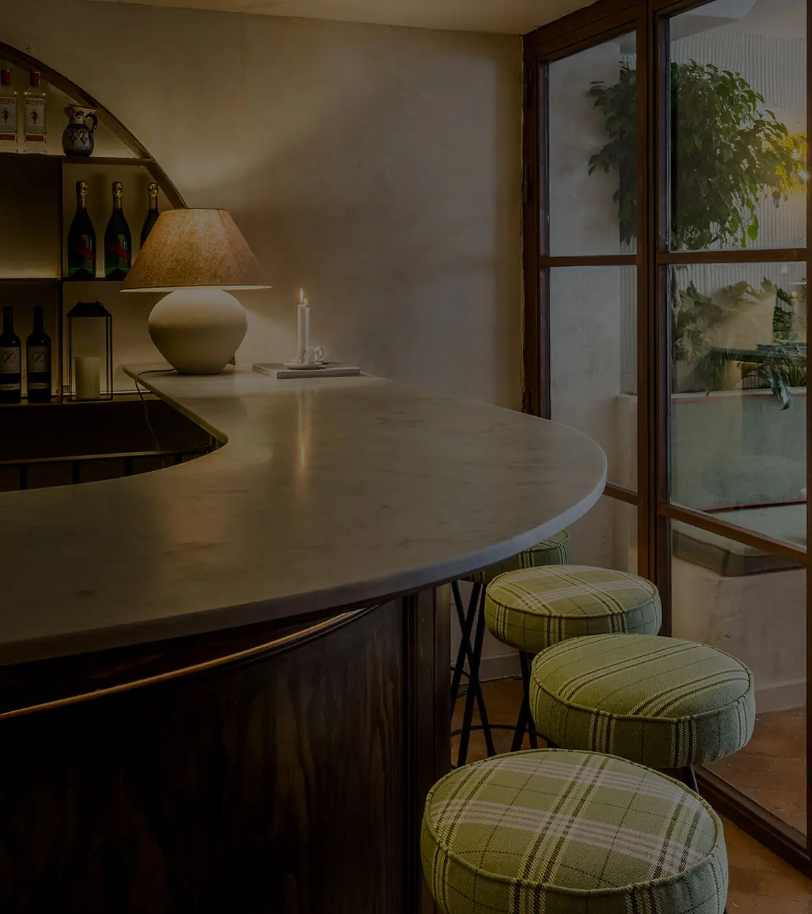 Corner of a curved marble bar with a ceramic table lamp, green plaid bar stools and a large window looking onto a plant-filled courtyard