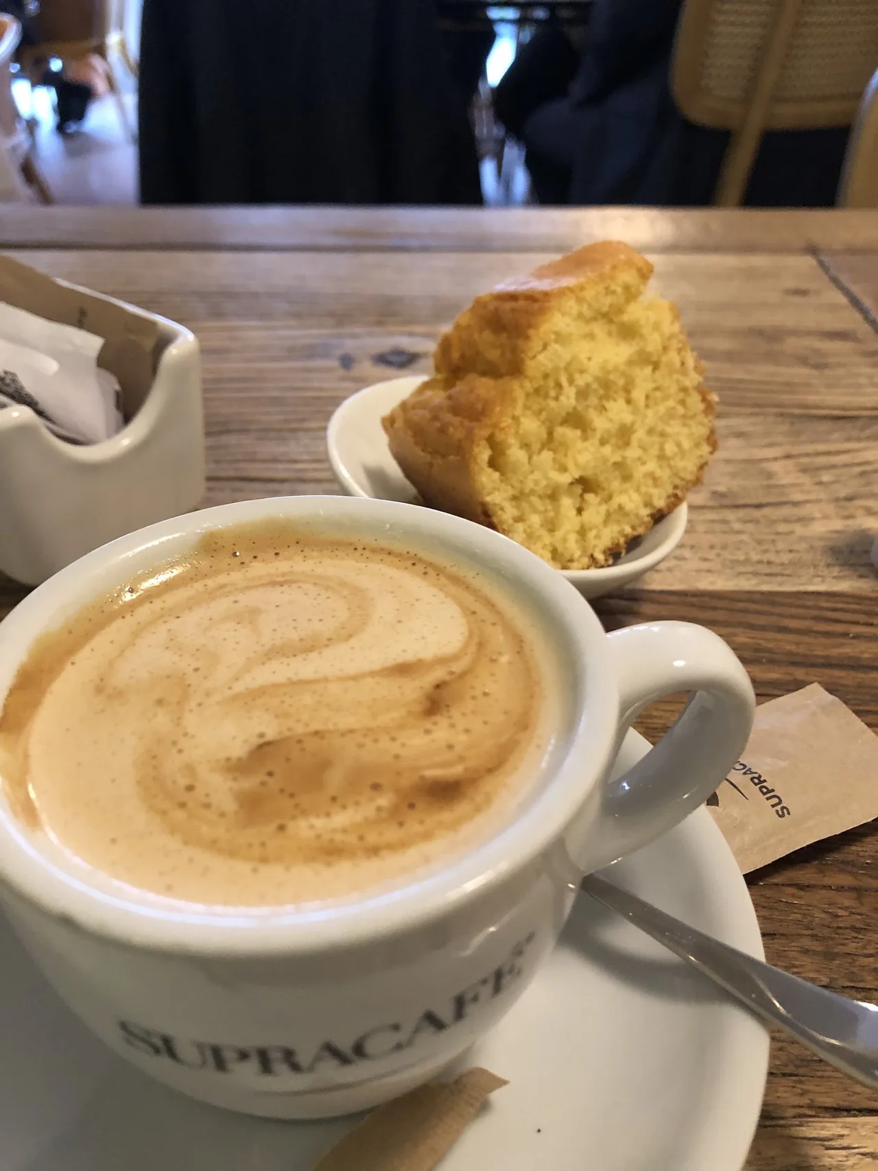 A cup of milky coffee with a swirl on top beside a slice of yellow sponge cake on a wooden table