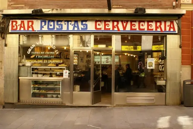 Exterior storefront of Bar Postas Cerveceria with red and blue signage, illuminated food display case in the window, and diners visible inside.