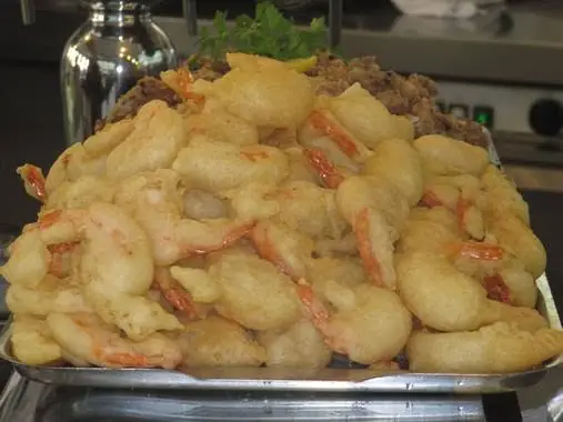 Metal tray piled with battered fried prawns, garnished with fresh herbs, on a stainless steel counter.