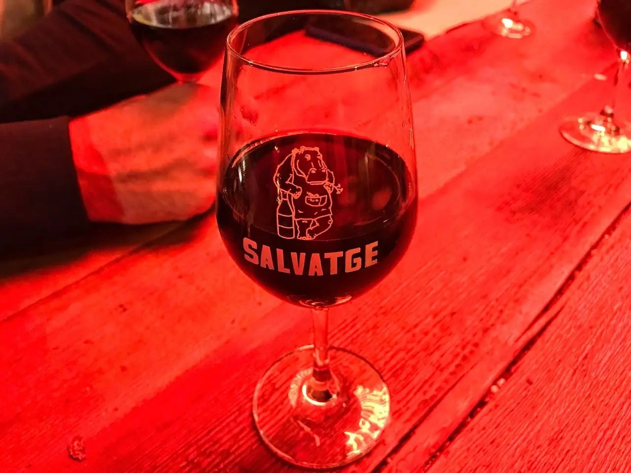 Glass of natural wine etched with the Salvatge logo on a wooden table at Bar Salvatge.