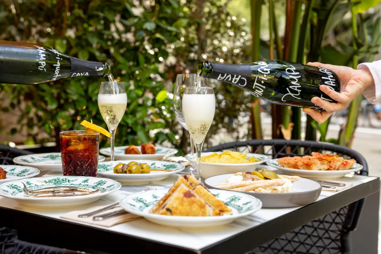 Cava being poured tableside alongside a spread of tapas plates — olives, anchovies, croquetas, cold cuts, and a Bloody Mary — on an outdoor table at Bar Vint-i-Quatre
