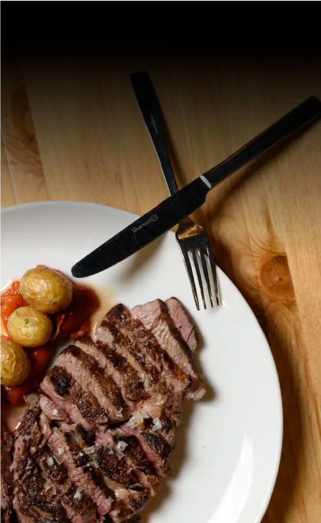 Sliced grilled steak with roasted baby potatoes and red pepper on a white plate with knife and fork