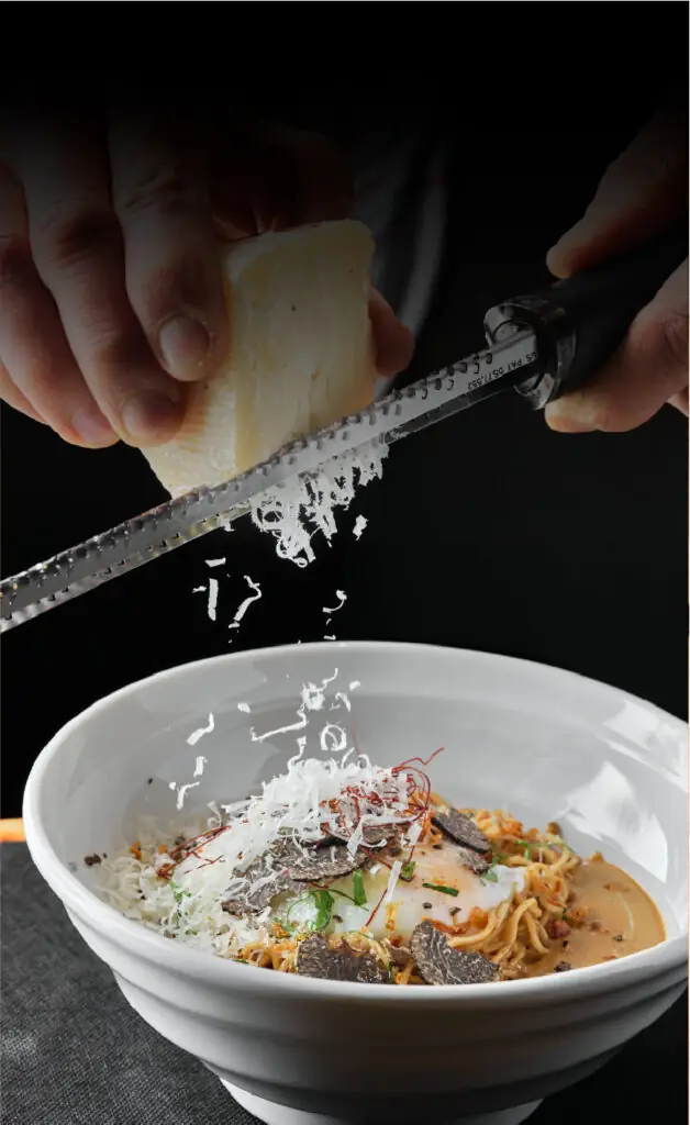 Hands grating hard cheese with a microplane over a bowl of noodles topped with truffle shavings and egg