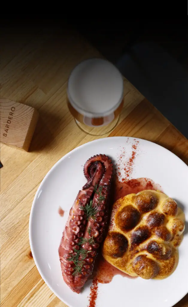Grilled octopus tentacle dusted with paprika beside a browned potato cloud on a white plate with a beer glass