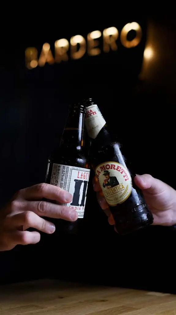 Two hands clinking Birra Moretti and Lagunitas beer bottles under an illuminated Bardero sign