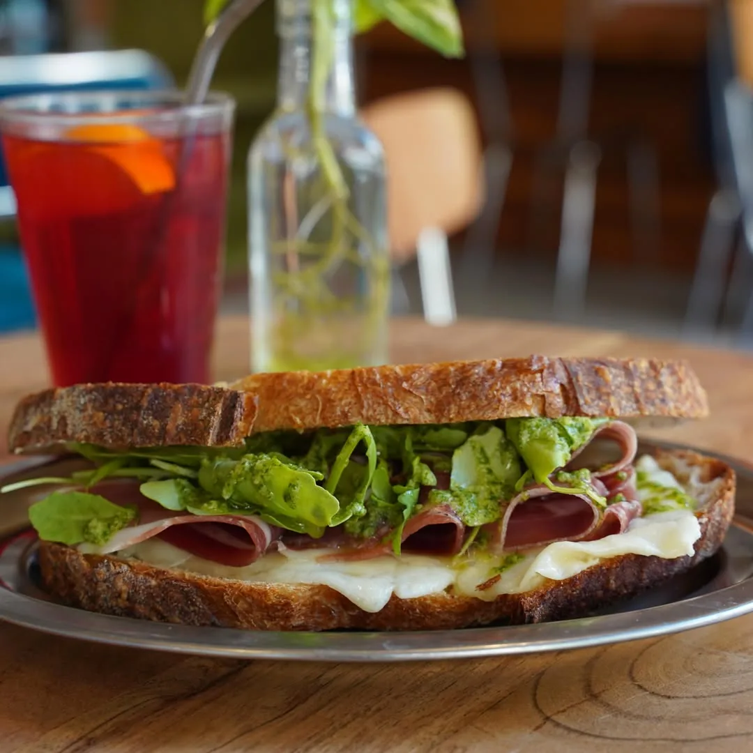 Thick sandwich with ham, cheese, and leafy greens on a plate, with an iced drink beside it