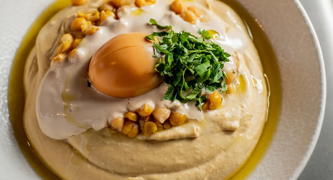 A close-up bowl of hummus swirled with olive oil, topped with tahini, whole chickpeas, a smooth orange quenelle and a heap of chopped parsley