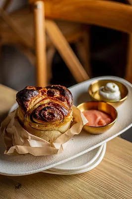 A glossy baked pastry roll on greaseproof paper on a pale oval plate, with two small brass dishes of pink sauce and white cream alongside, on a wooden table