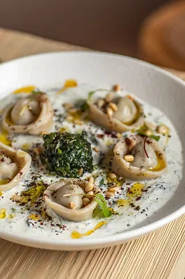 Filled dumplings sitting in a pool of yoghurt in a white bowl, dressed with olive oil, sumac, toasted pine nuts and a spoonful of dark green herb paste at the centre