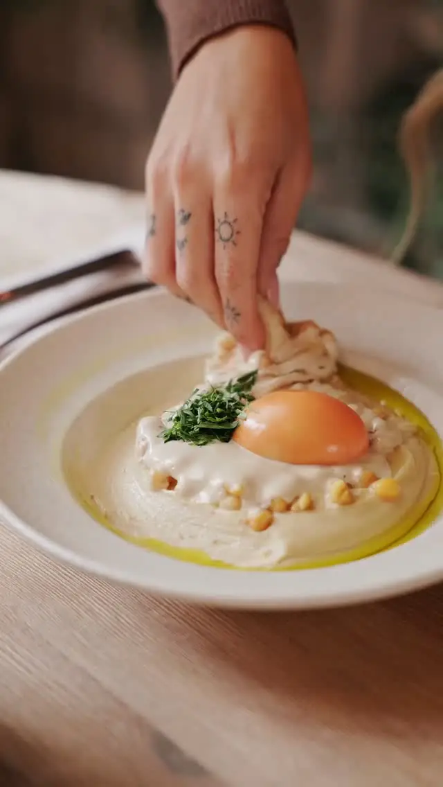 Hand draping tahini sauce over a creamy dip topped with a soft egg yolk and herbs