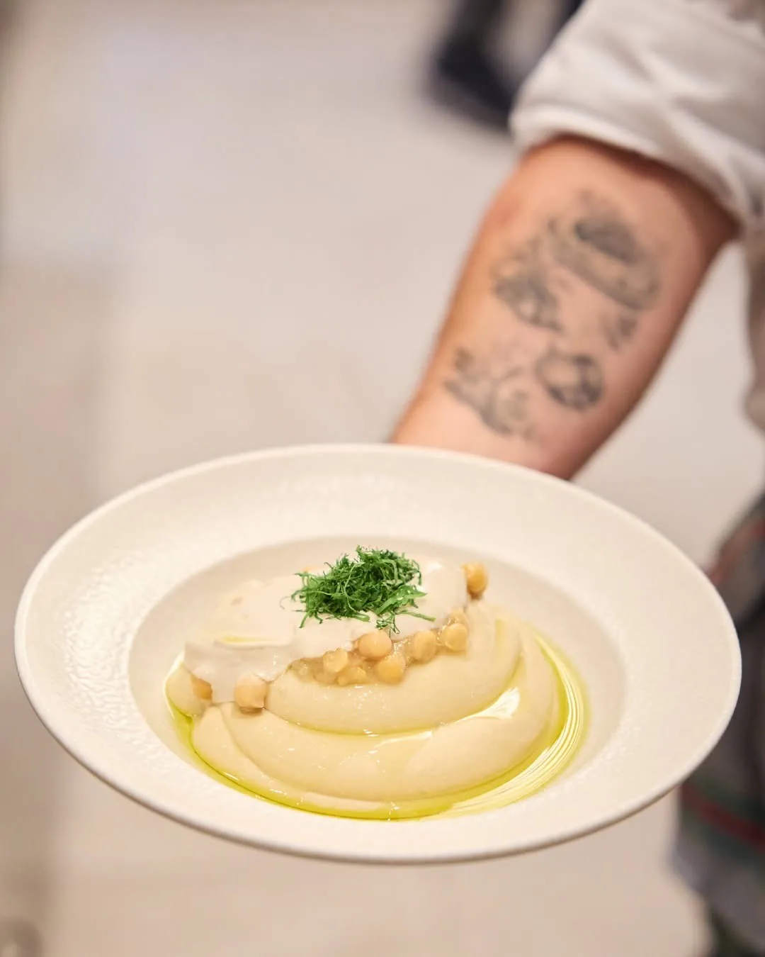 A bowl of creamy hummus topped with chickpeas and herbs, held by a hand with a tattooed arm