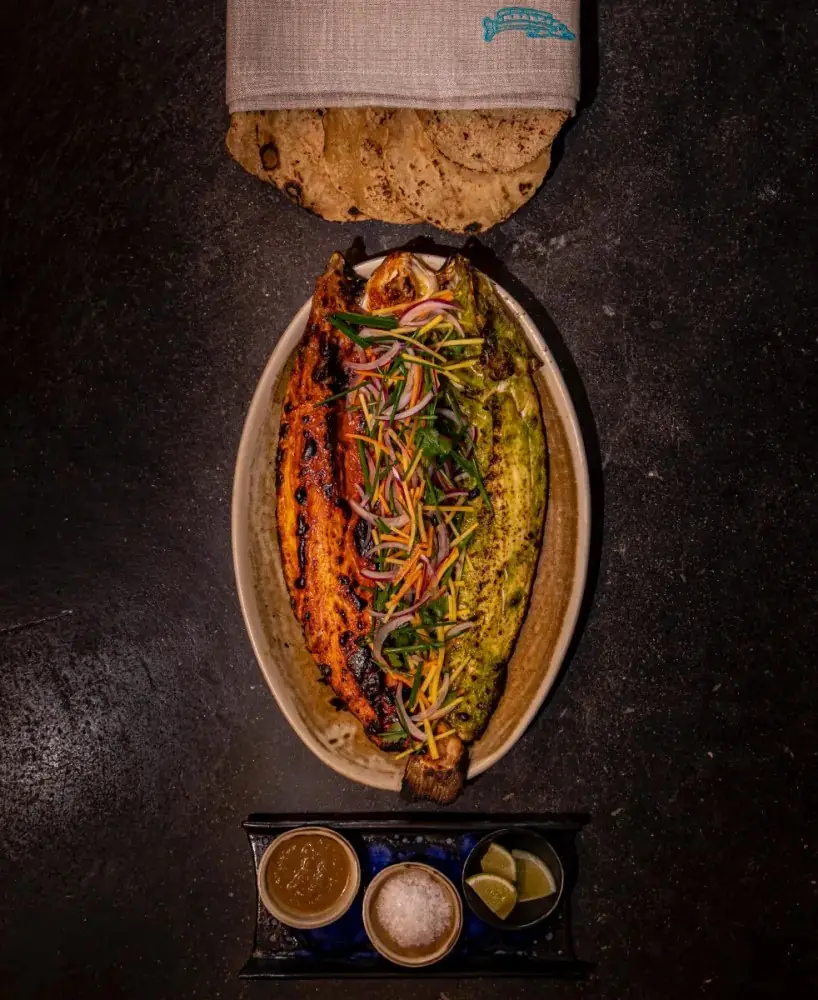 A whole butterflied grilled fish on an oval stone plate, one half glazed red and one half green, heaped with coriander and shredded red onion and carrot, with a cloth-wrapped stack of tortillas above and a tray of salsa, salt and lime wedges below