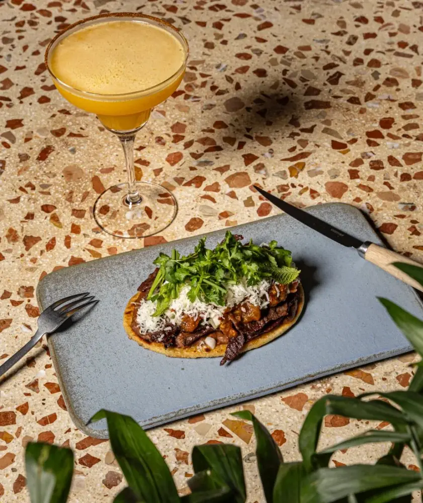 A tostada piled with strips of dark meat, orange sauce, grated white cheese and fresh coriander on a grey slate board, with a foamy yellow cocktail in a coupe behind, on a terrazzo table