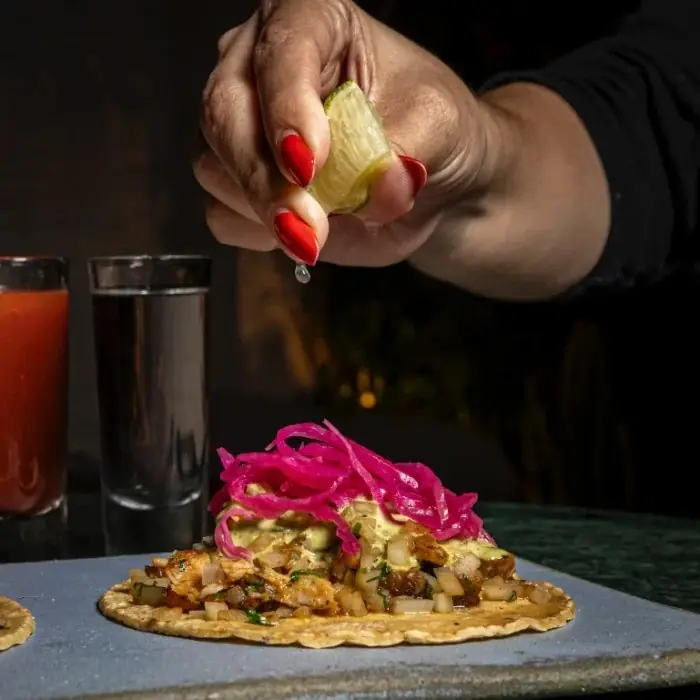 A hand with red nails squeezing a lime wedge over a taco of chopped seafood, onion and yellow sauce topped with pickled pink onion, with a red drink and a shot glass behind