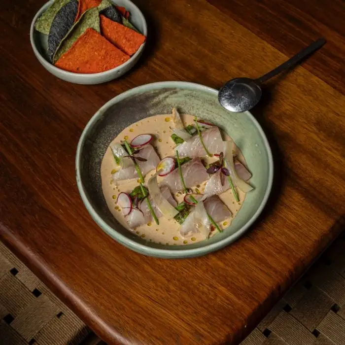 A pale green bowl of raw white fish slices in a creamy tan sauce dotted with oil, garnished with radish slices and chives, beside a small bowl of blue and red tortilla chips on a dark wood table