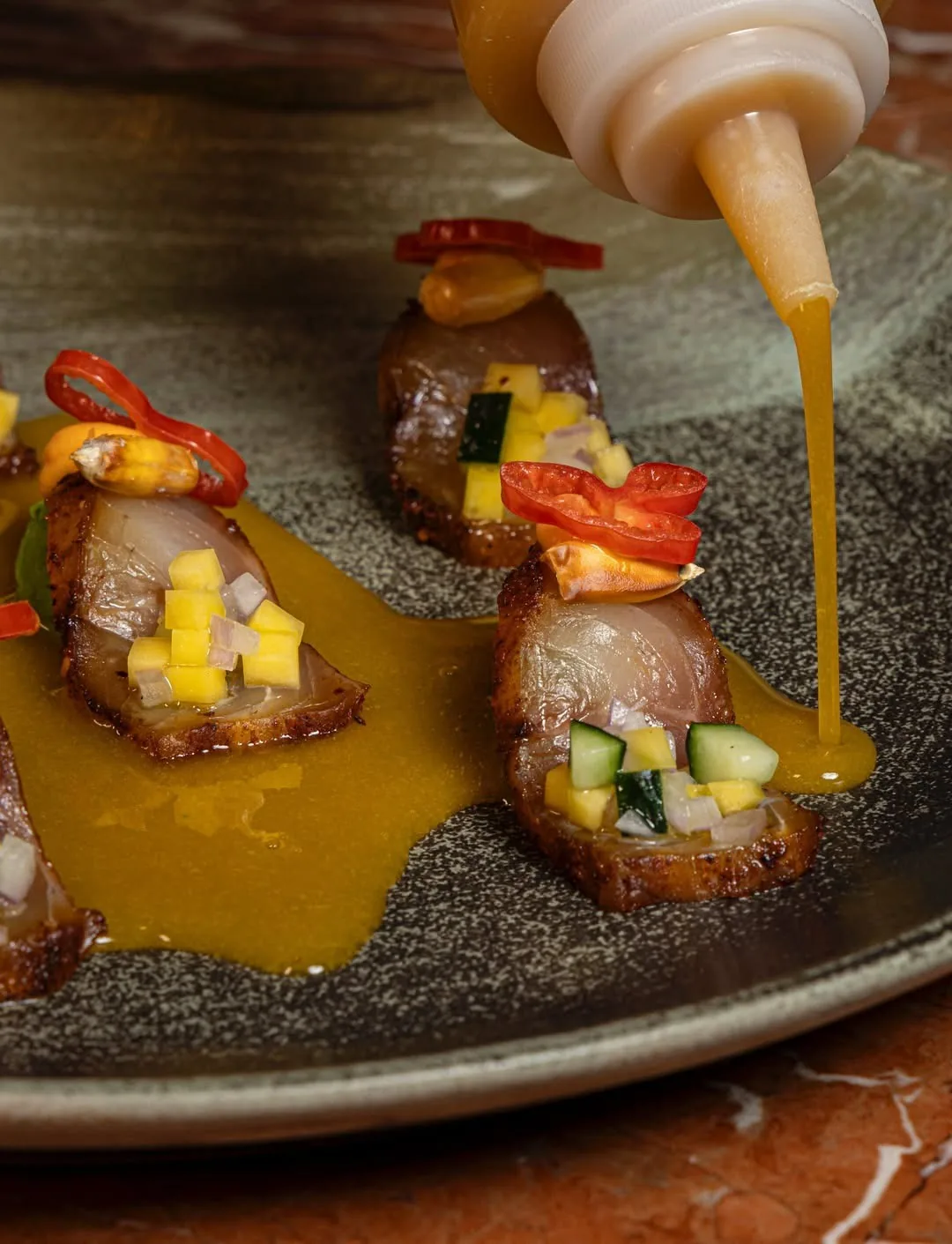 A dark plate with three small dishes topped with diced vegetables and drizzled with yellow sauce