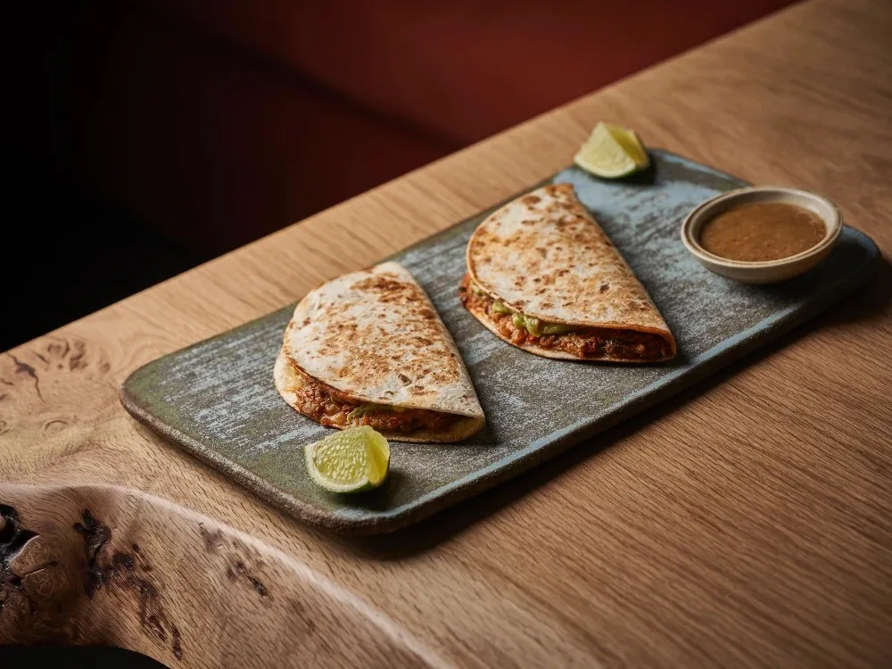 Two folded golden quesadillas on a slate tray with lime wedges and a small bowl of sauce