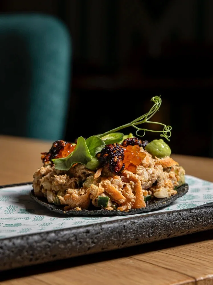A dark round tostada topped with a chopped mix and small green leaves on a grey plate