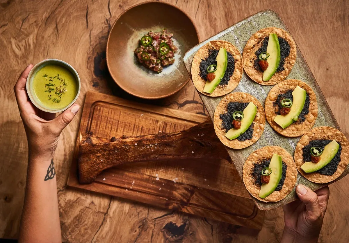 A wooden board with small round crisp tortillas topped with avocado, beside a green bowl held by a hand