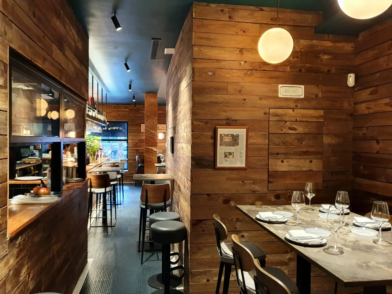 Narrow tapas bar clad in reclaimed timber, with a kitchen hatch and bar stools along the left, a globe pendant lamp, a framed newspaper article on the wall and a marble table laid with wine glasses and napkins
