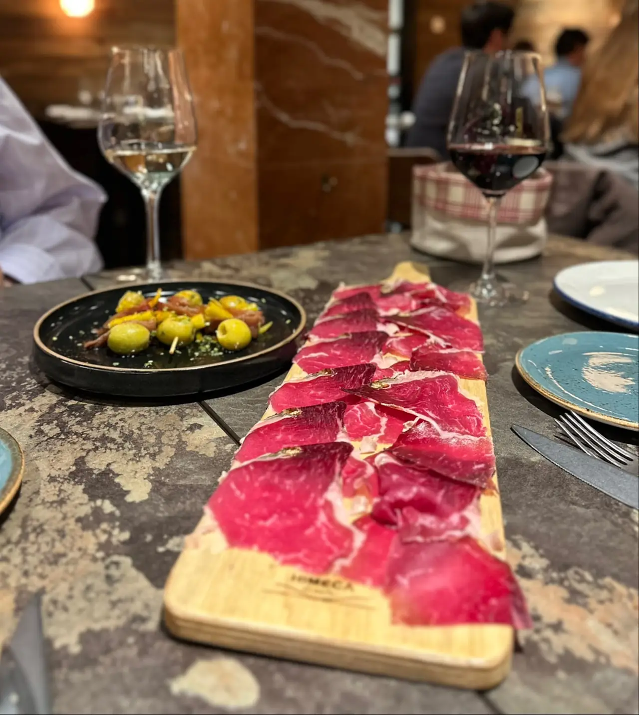 Sliced cured meat on a wooden board beside a plate of olive and anchovy skewers