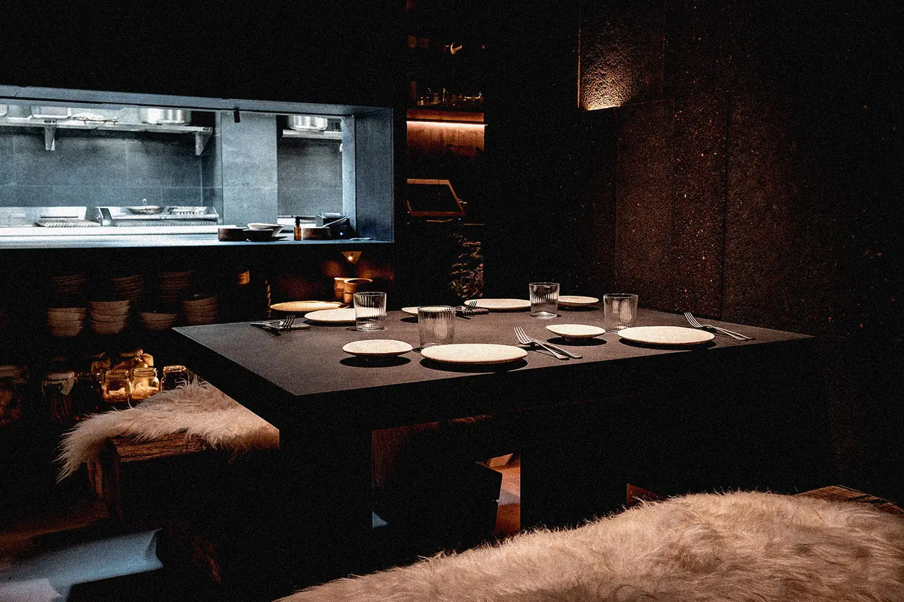 A dark dining room: a black table laid with pale stoneware plates, ribbed tumblers and cutlery, sheepskin fleeces draped over the benches, shelves of stacked bowls and glass preserve jars at the left, and a lit open-kitchen pass window behind.