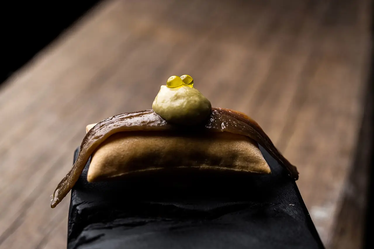 A cured anchovy fillet draped over a puffed golden crisp on a black slate, topped with a pale green quenelle crowned by two olive-oil pearls, wooden counter behind.