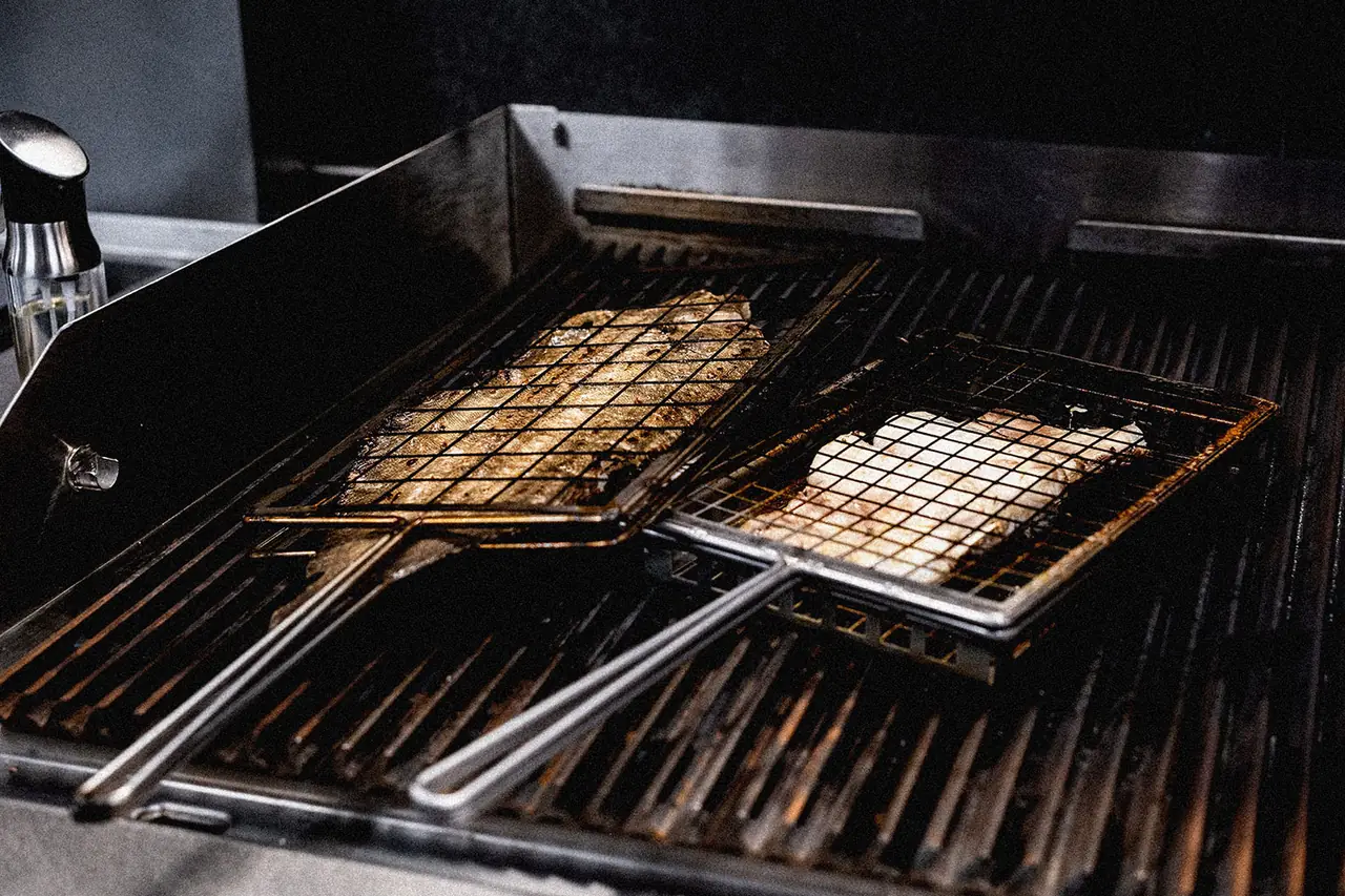 Two hinged wire grilling baskets on a charcoal grill: one holding grilled sardines, the other a thick white fish fillet, with an oil dispenser at the edge.