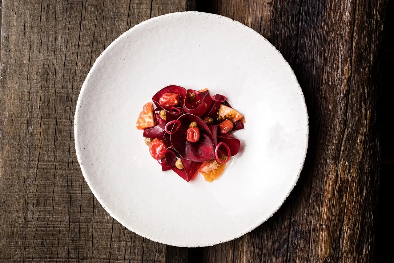 Overhead of a white speckled ceramic bowl on weathered wood holding thin folded sheets of beetroot with tomato wedges, pickled red onion rings and capers.