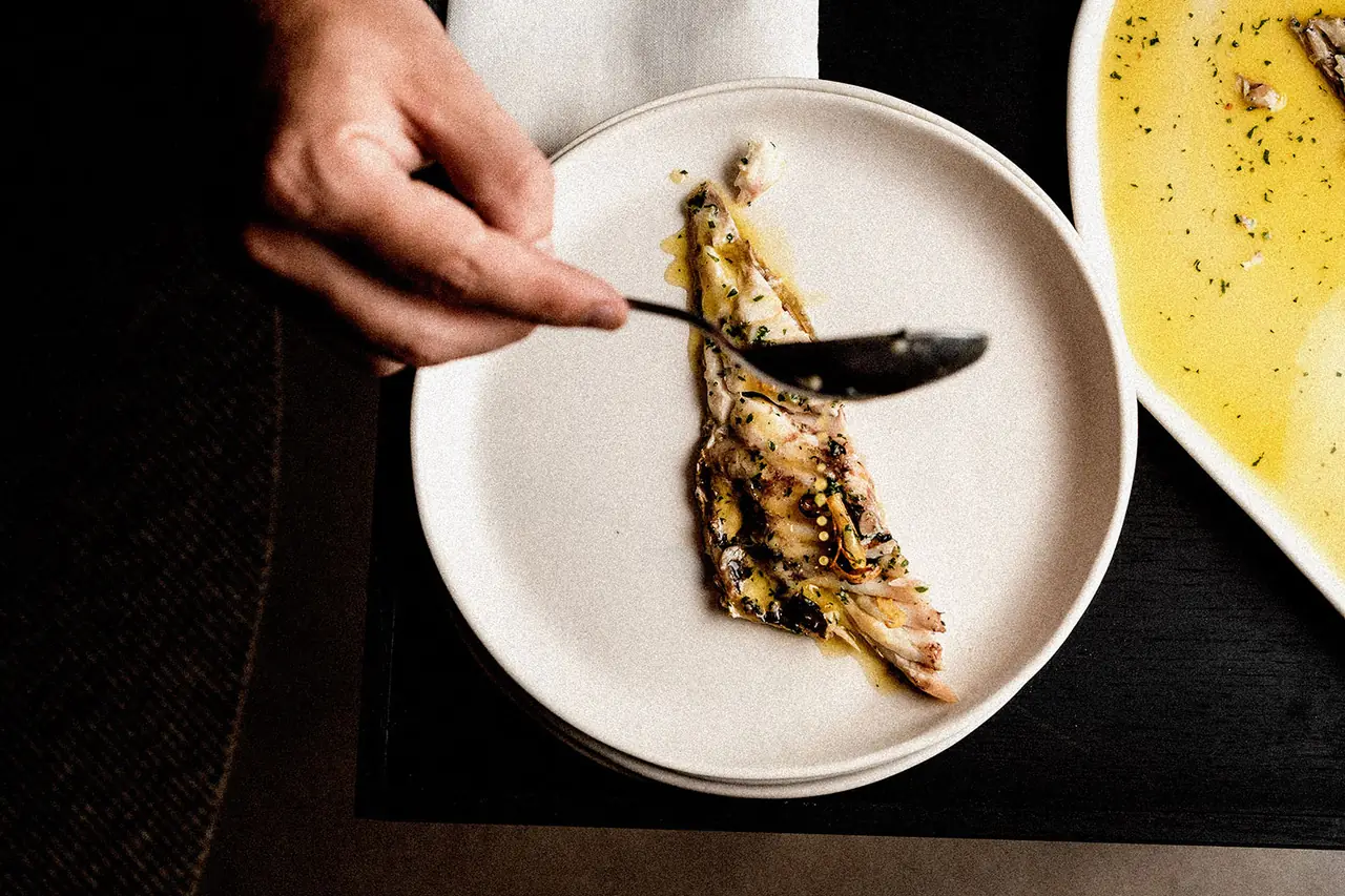 Hand plating a grilled fish fillet with herb oil onto a white plate, another dish of yellow sauce beside it