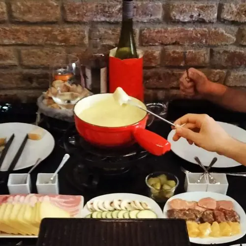 Cheese fondue pot with a fork dipping bread, served with charcuterie, sliced zucchini, mushrooms, ham, and pineapple at Basilea Restaurant