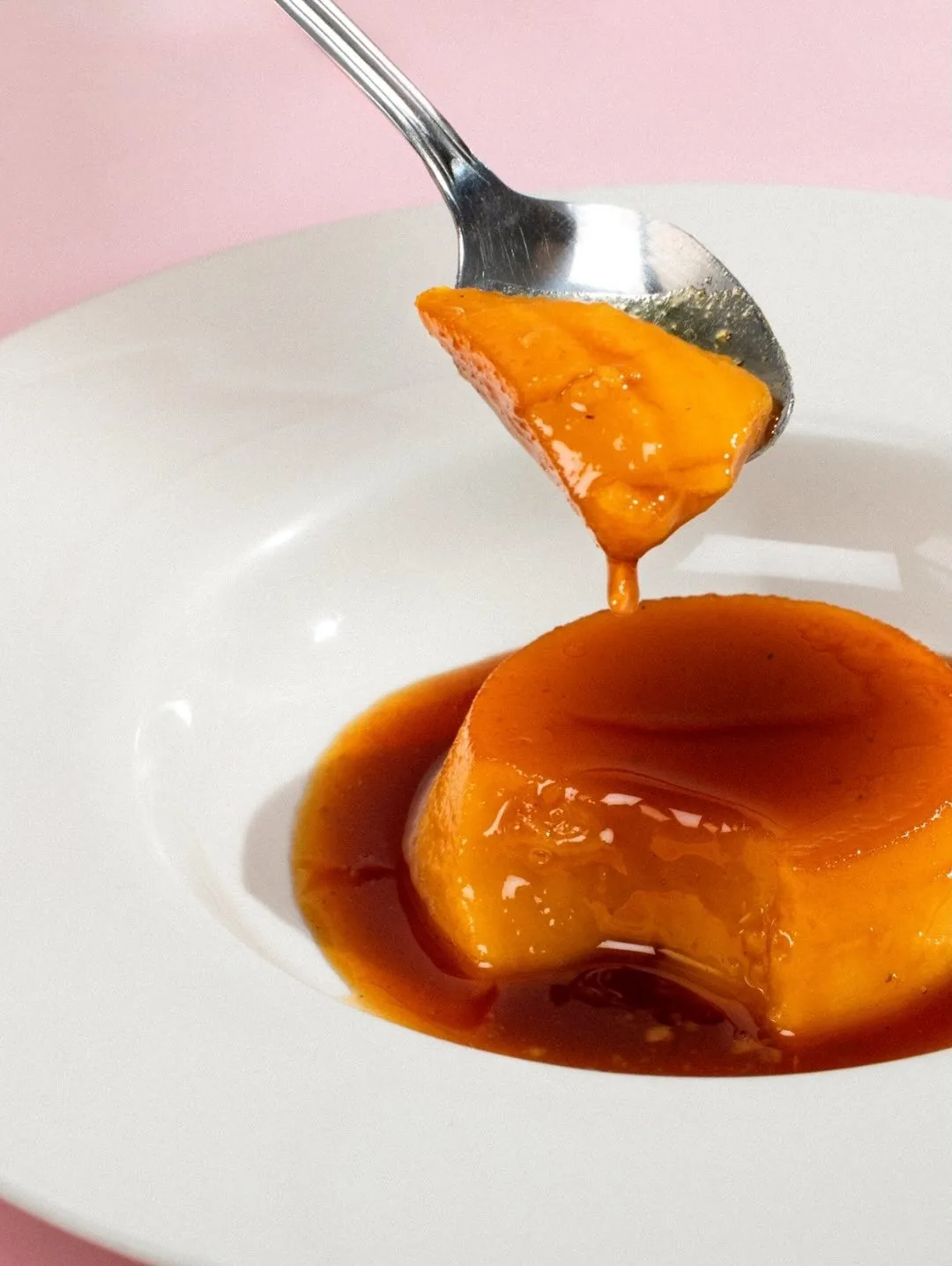 A round caramel-topped custard being drizzled with orange sauce on a white plate